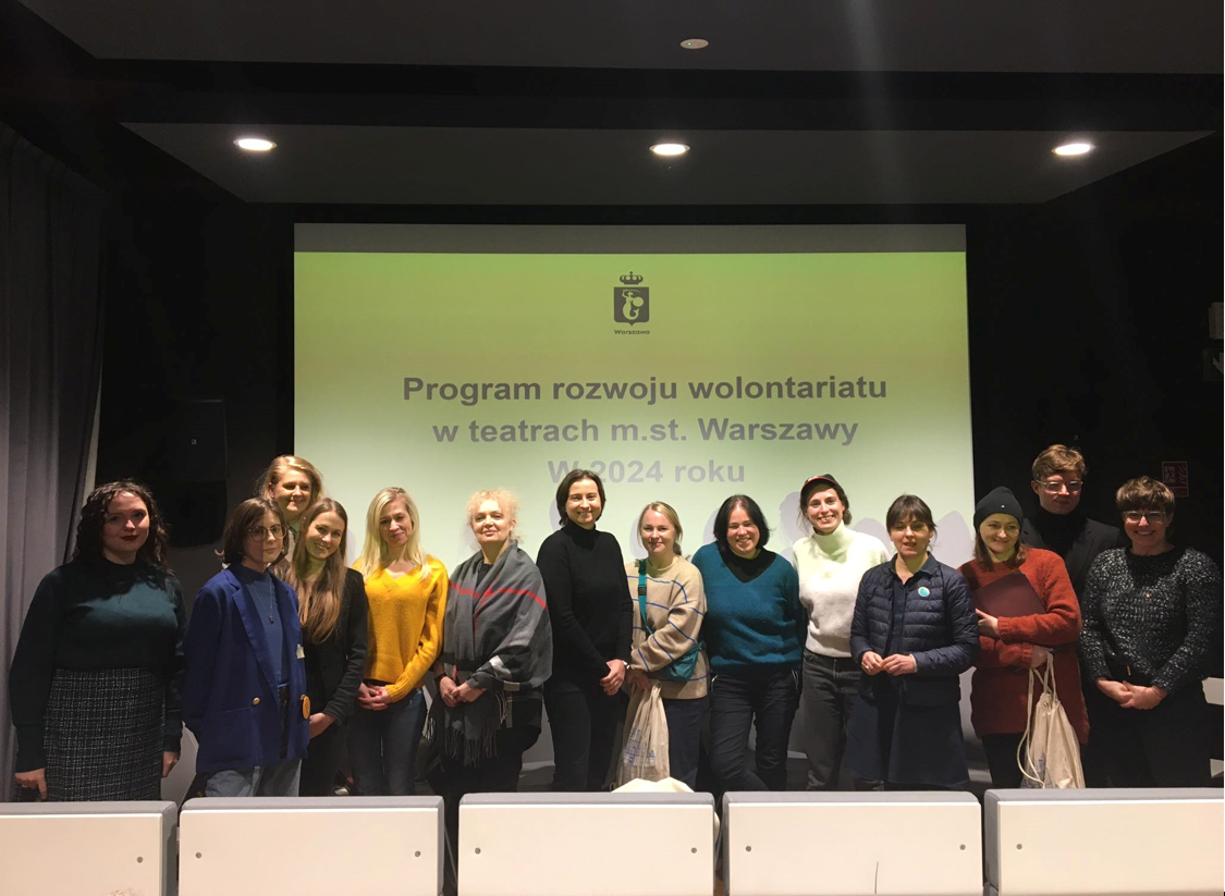 Grupa ludzi stojących przed żółtym ekranem z napisem "Program rozwoju wolontariatu w teatrach m.st. Warszawy w 2024 roku"