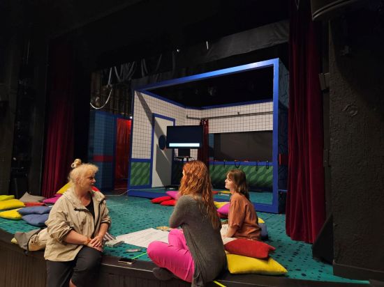 Kilka osób siedzi na kolorowych poduszkach na scenie teatralnej i rozmawia, scena jest zielona, w tle czerwona kurtyna i scenografia o tematyce sportowej