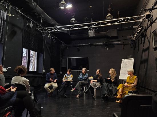 Kilka osób siedzi w kręgu na krzesłach w sali teatralnej i dyskutuje, sala jest ciemna - z czarną podłogą i czarnymi ścianami, wyposażone w duże reflektory teatralne