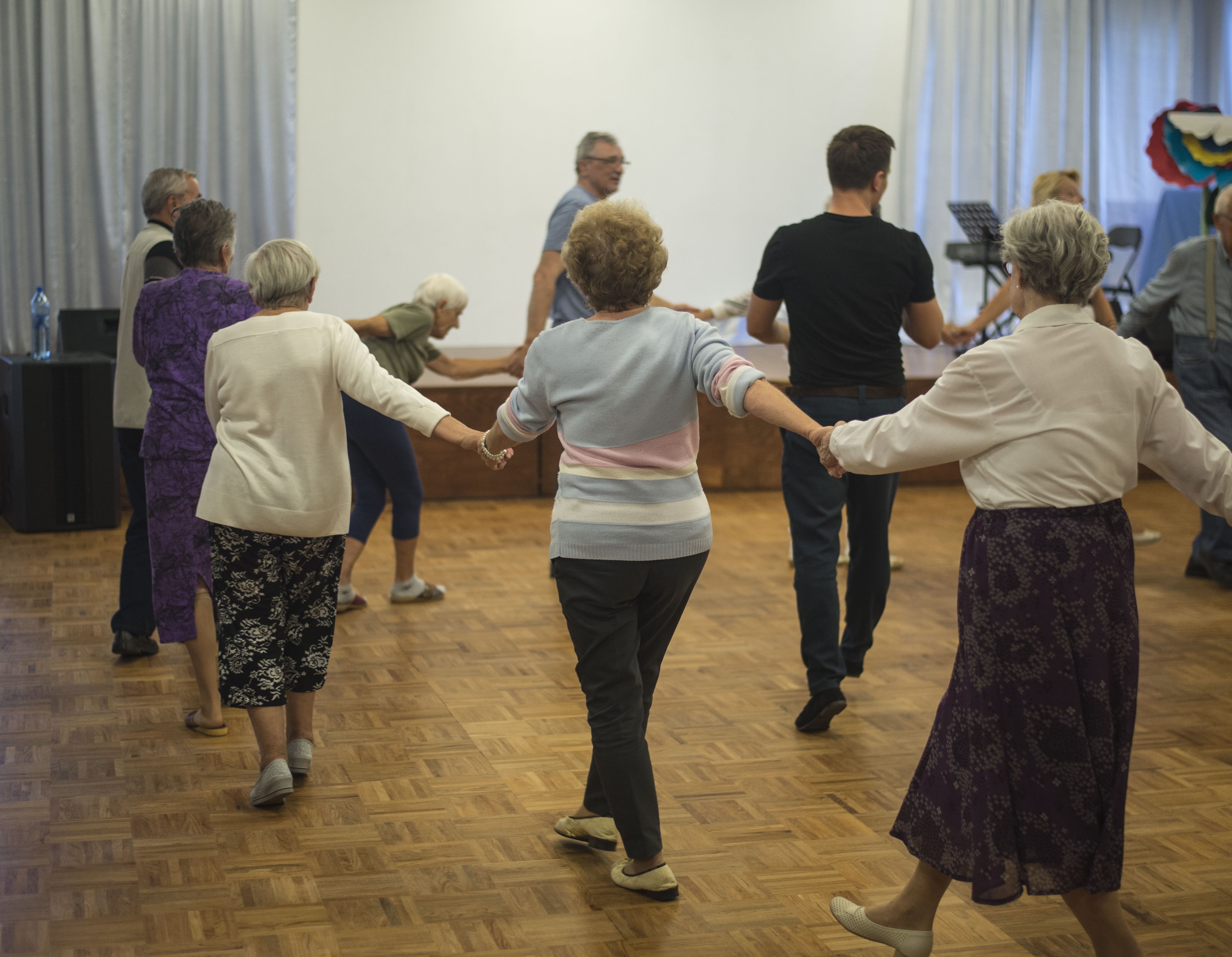 Seniorzy trzymają się za ręce i tańczą w kole podczas zajęć tanecznych