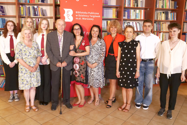 Pracowniczki biblioteki wraz z młodzieżą i powstańcami warszawskimi pozują do zdjęcia. Za nimi znajduje się regal z książkami.