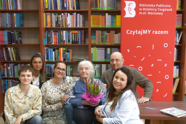 Pracowniczki i pracownicy biblioteki pozujący do zdjęcia wraz panią Joanną Papuzińską. Za nimi znajduje się regał z książkami.