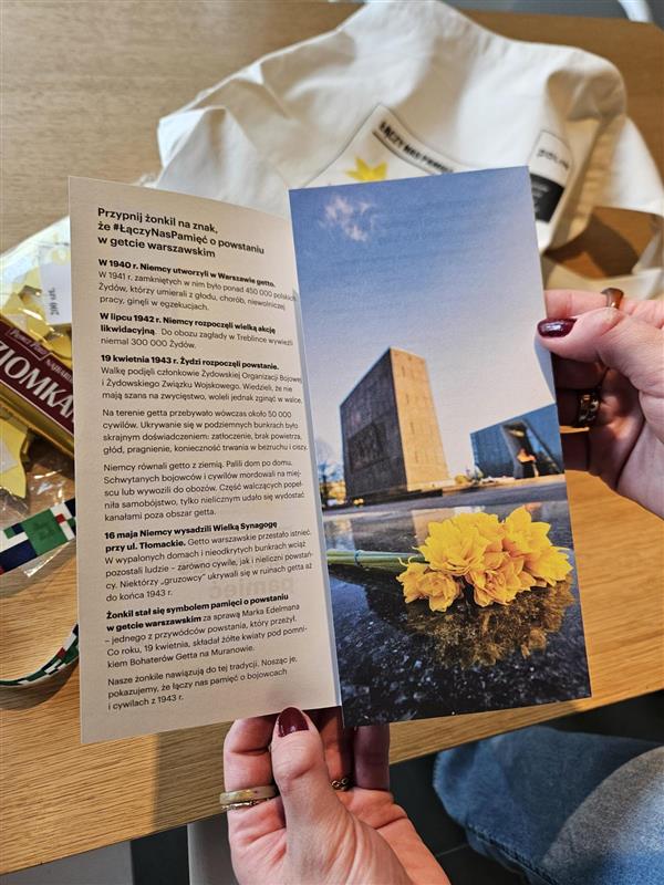 Ręka trzymająca otwartą ulotkę. Po lewej stronie widnieje czarny napis "przypnij żonkil na znak, że #łączy nas pamięć". Po prawej stronie widać zdjęcie leżącego bukietu żonkili.