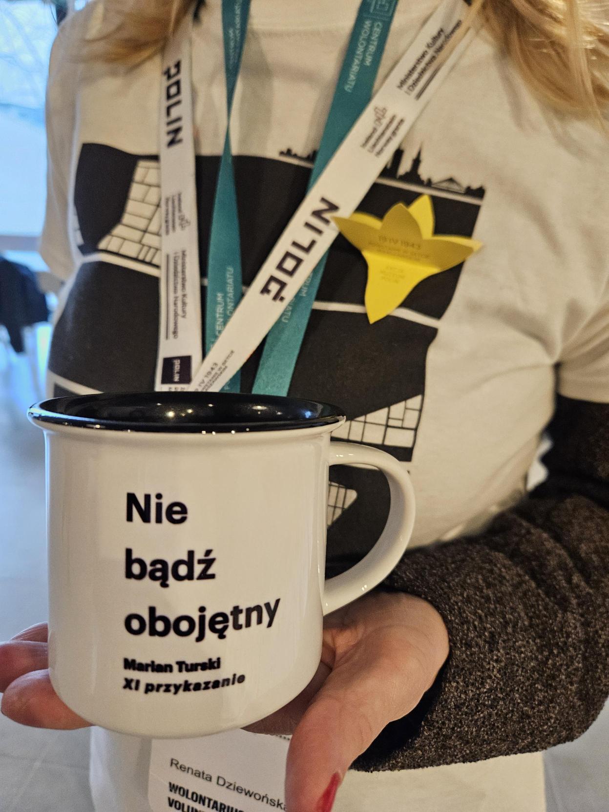 Ręka osoby trzymającej aluminiowy biały kubek z czarnym napisem "nie bądź obojętny"