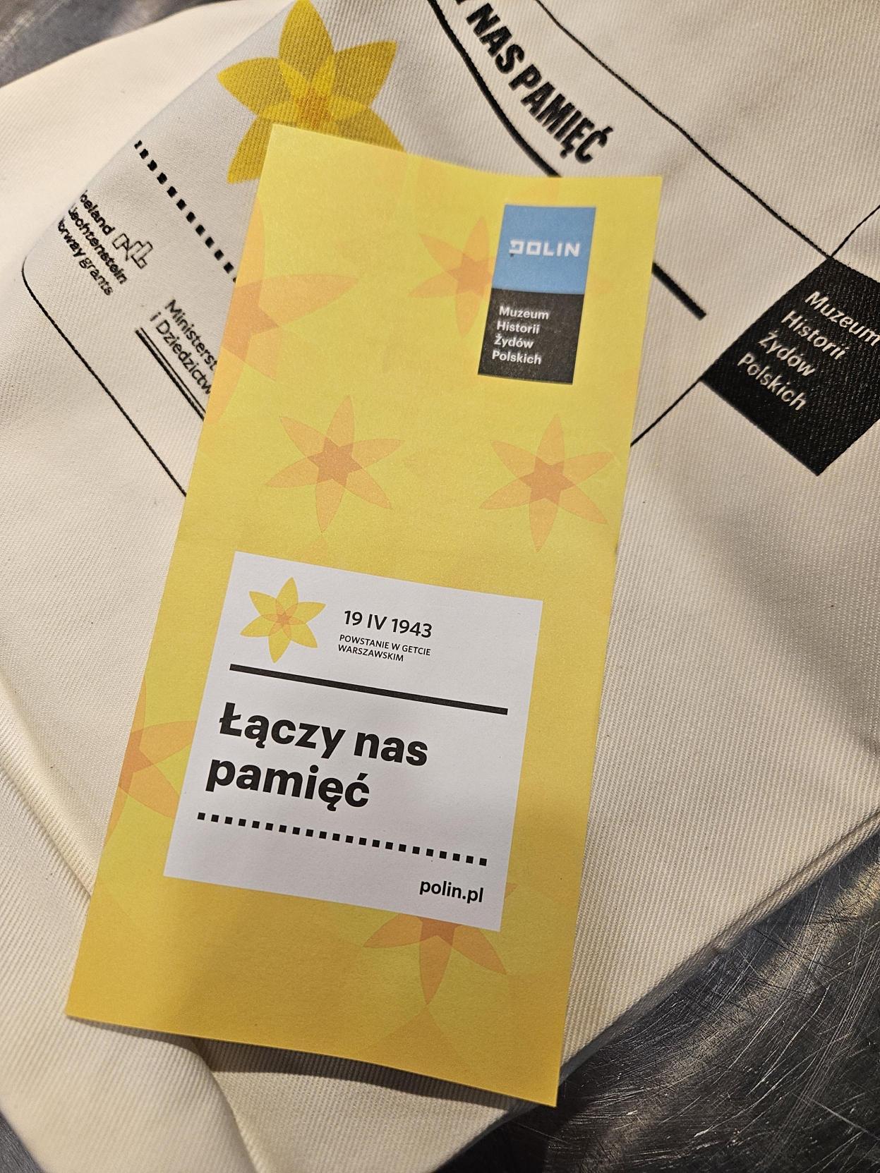 Żółta ulotka w żonkile z czarnym napisem "łączy nas pamięć" umieszczonym w białej ramce.