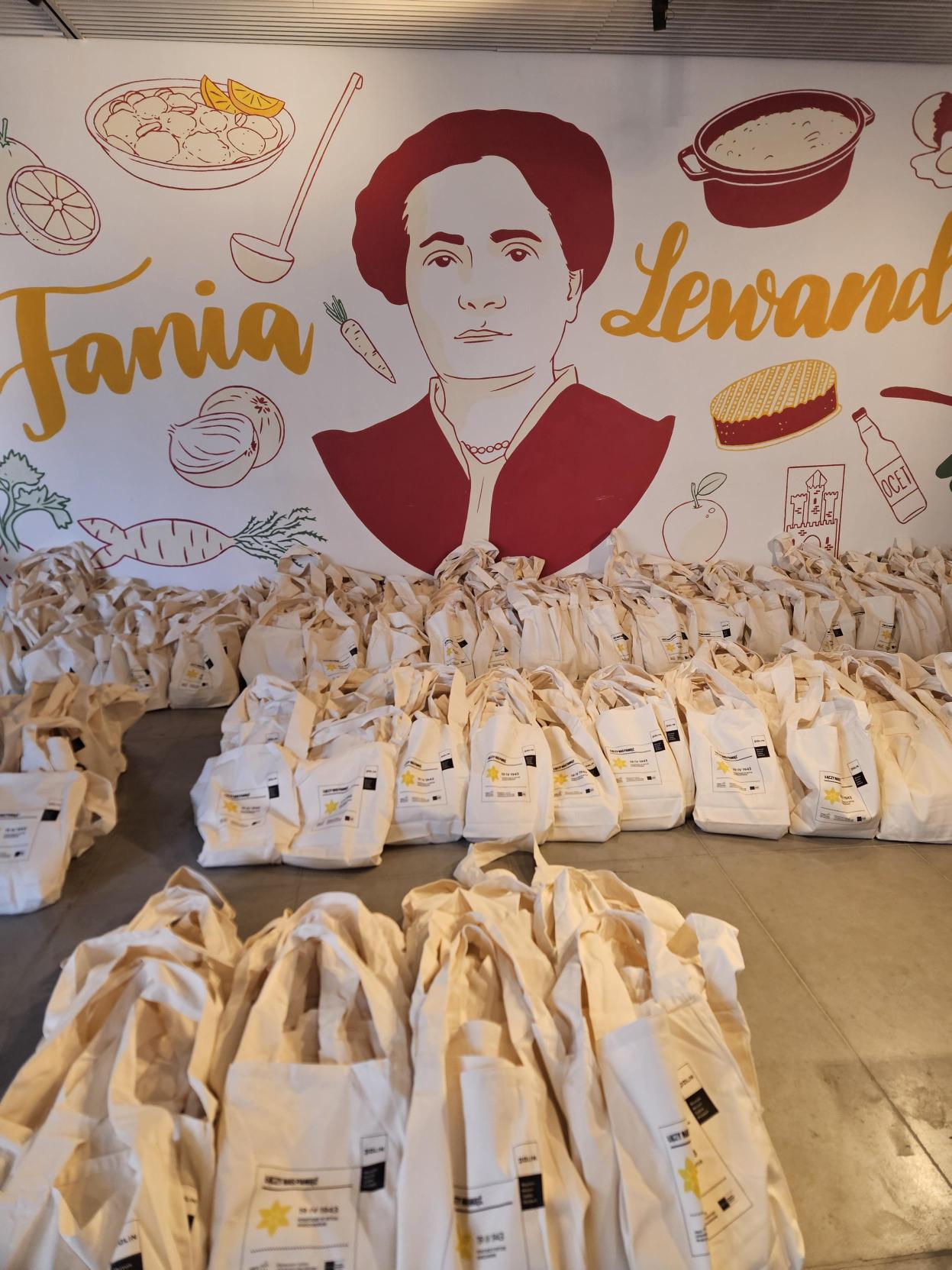 Bawełniane torby z wizerunkiem żonkili leżące na podłodze. Za nimi widać ścianę z muralem przedstawiającym kobietę oraz podpisem "Fania Lewando"