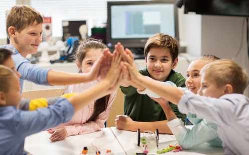 Zdjęcie siedmiorga dzieci w wieku wczesnoszkolnym siedzących w sali dookoła stolika, wyciągających przed siebie rękę, łącząc dłonie 