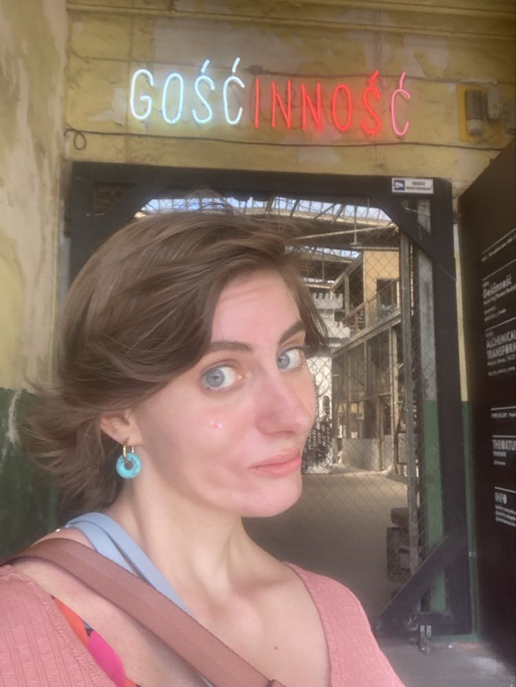 Zdjęcie portretowe kobiety stojącej na tle neonowego biało-czerwonego napisu "GOŚĆ-INNOŚĆ". Kobieta ma krótkie brązowe włosy, turkusowe kolczyki i jest ubrana w łososiową bluzkę.  