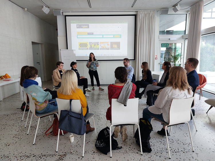 Kilkunastu uczestników siedzących na białych krzesłach w kręgu, słuchających prowadzącej