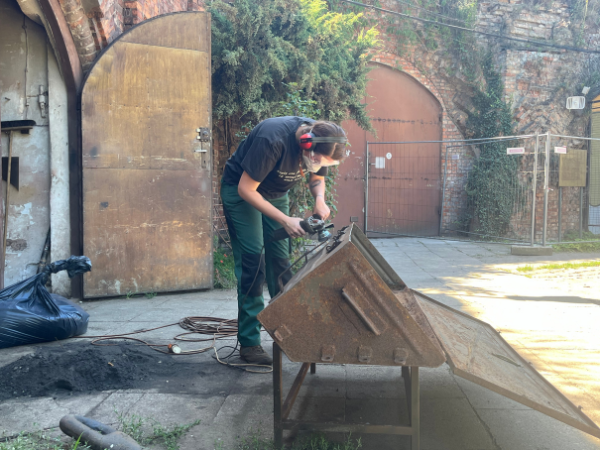 Kobieta ubrana w czarną koszulkę oraz butelkowo-zielonych spodniach pochylająca się ze spawarką nad metalowym fragmentem