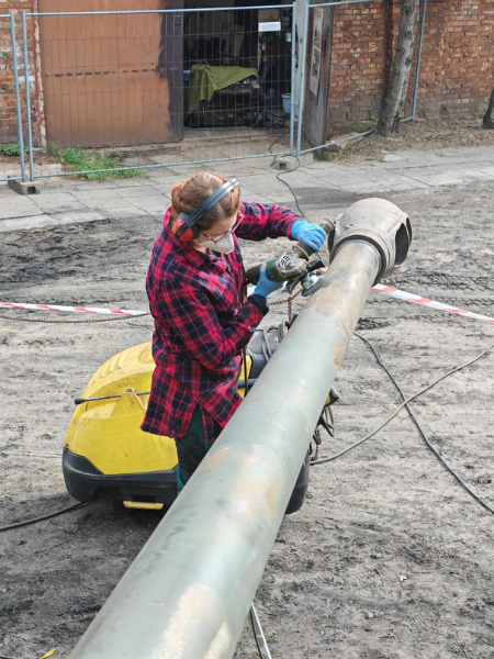Kobieta ubrana w koszulę w czerwono-czarną kratę, słuchawki ochronne oraz niebiskie rękawiczki. W ręku trzyma szlifierkę, którą dotyka lufy czołgu