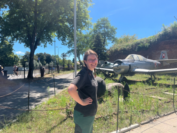 Kobieta ubrana w czarną koszulkę oraz ciemnozielone spodnie. Stoi bokiem, z ręką na biodrze i się uśmiecha. Dookoła niej widać wojskowe eksponaty ustawione na zielonym terenie