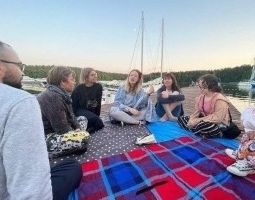 Grupa ludzi siedzących na kocach w kręgu na kei, na pierwszym planie jaskrawy koc w czerwoną i szafirową kratę, w tle jezioro i wystające maszty zacumowanych żaglówek