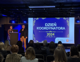 Zdjęcie przedstawia scenę na której stoi Antoni Morawski, kierownik Zespołu Rozwoju Wolontariatu w Centrum Komunikacji Społecznej oraz Agnieszka Lissowska-Lewkowicz, prezes Stowarzyszenia Centrum Wolontariatu. Z tyły wyświetlona jest grafika z napisem "Dzień Koordynatora 2024"
